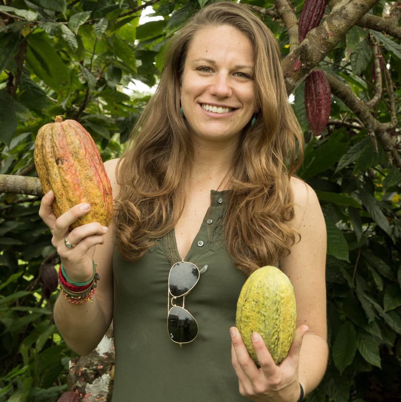 A woman smiling while holding cacao pods in front of trees