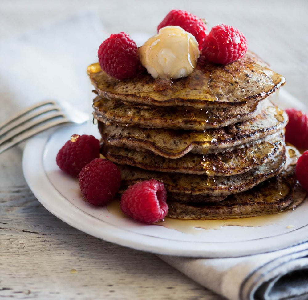 a stack of cacao pancakes topped with maple syrup, raspberries, and vegan butter